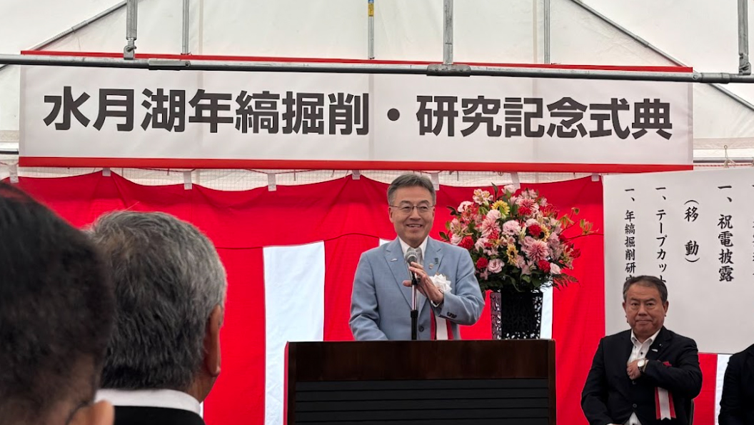 「祝 水月湖年縞掘削・研究記念式典」と書かれた吊り看板の下で笑顔で挨拶する男性