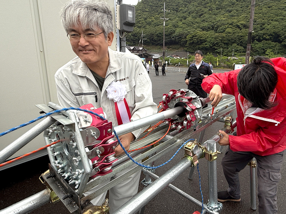 長いパイプの周りに金具がたくさんついた装置の前で笑顔を見せる男性