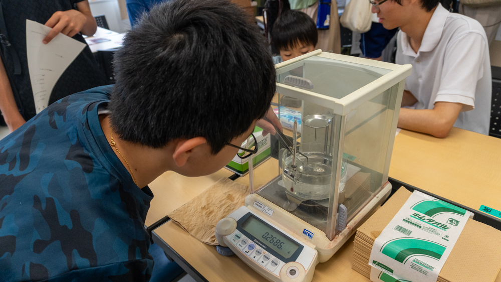 水が入ったビーカーが設置された電子天秤のかごにピンセットで試料を載せる少年のようす