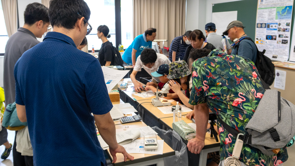 それぞれの机の上に置かれた実験装置をのぞき込む人たちのようす