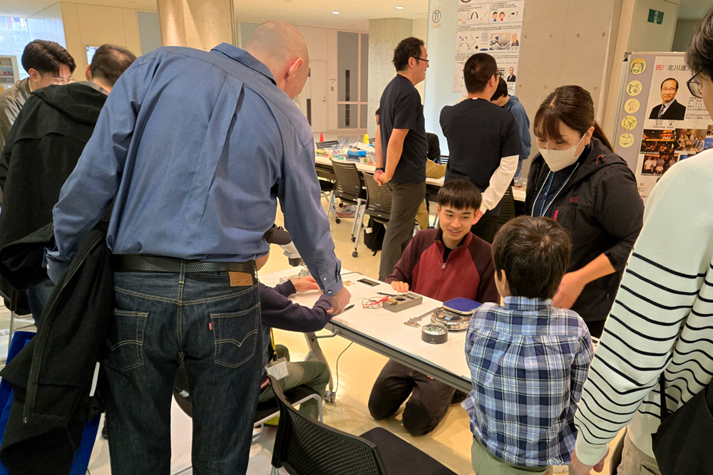 テスターや顕微鏡などが置かれたテーブルで実験する子どもたちと笑顔で対応するスタッフ