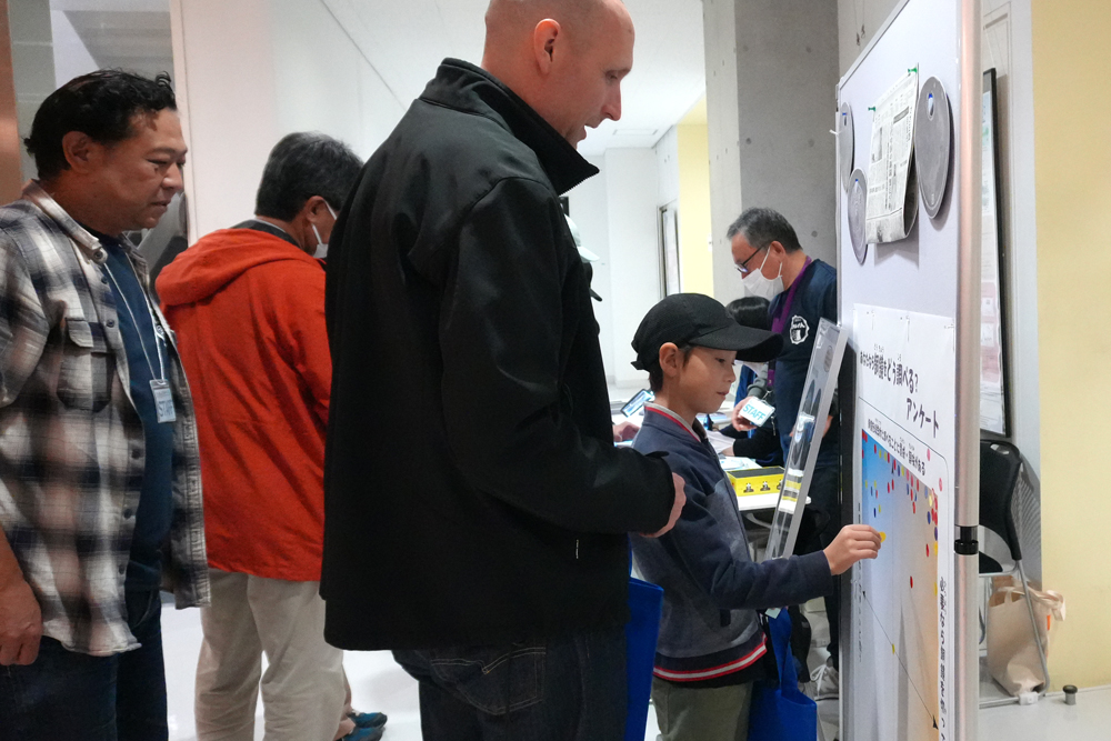 ボードに貼られたアンケート用チャートに小さなシールを貼る親子のようす