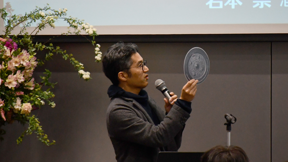 丸い紙うちわを見せながら話す男性