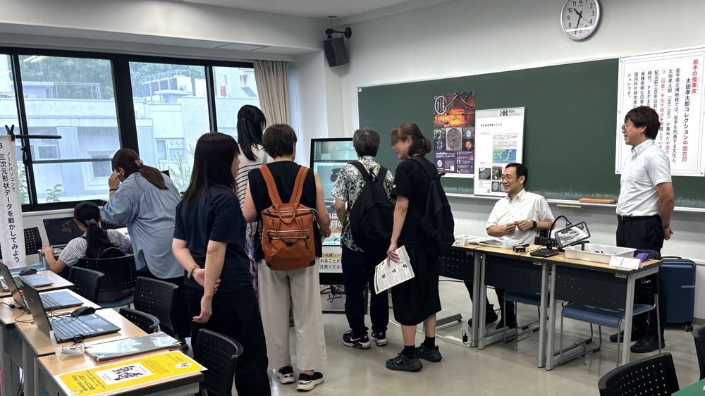教室の一角に置かれた縦型モニターに映る動画を見る人々とそれを見ている人々