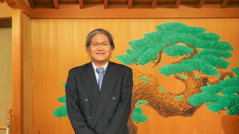 松が描かれた能楽堂を背景にした男性の写真