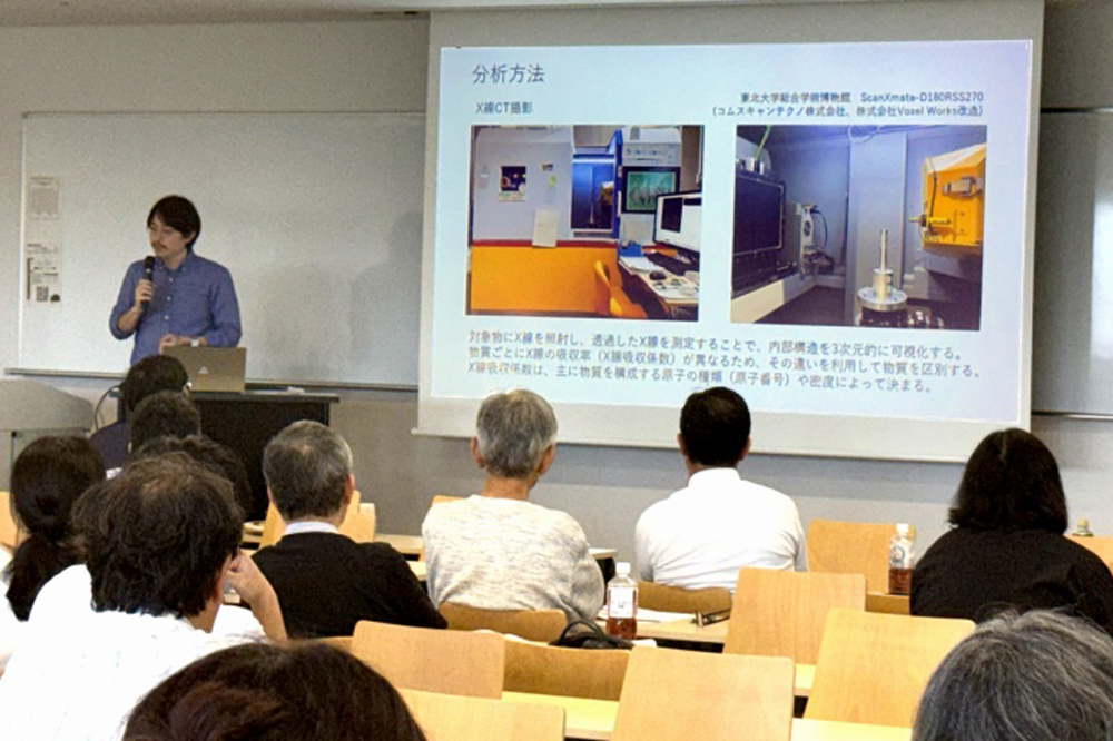 スクリーンの横でマイクを持って話している男性