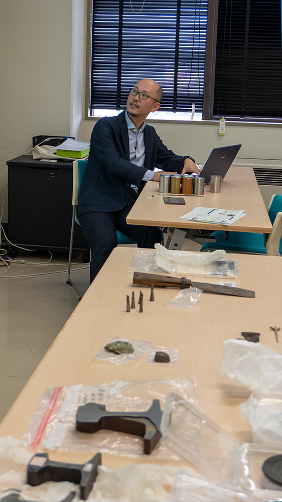 PCを開いている男性と、多数の金属製品が並ぶ机
